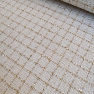 Puede incluir: Una tela texturizada de color blanco roto que presenta un patrón de cuadrícula repetitivo creado con hilo dorado metálico. El material tiene una apariencia tejida con fibras visibles y un diseño de cuadros cuadrados consistente en toda la superficie.