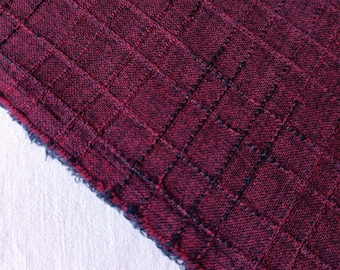 May include: A piece of deep burgundy fabric featuring a textured grid pattern created by dark black woven lines. The material has a visible weave structure with a slightly frayed, raw edge along the diagonal cut, showing the dark thread density.