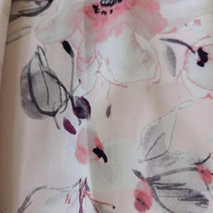 May include: Close-up of a fabric with a floral pattern. The design features large, stylized flowers in shades of pink, white, and black against a pale pink background. The leaves are a muted gray. The fabric appears to be soft and flowing.