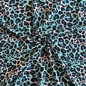 Puede incluir: Una tela con un estampado de leopardo en color turquesa y negro. La tela tiene un fondo blanco con manchas turquesas y negras. La tela está doblada y drapeada, mostrando la textura y el patrón del material.