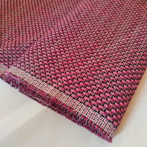 May include: A folded piece of woven fabric featuring a dense, textured pattern of vibrant magenta and black threads. The material displays a distinct grid-like weave structure with a visible, light-colored selvage edge along the folded corner.