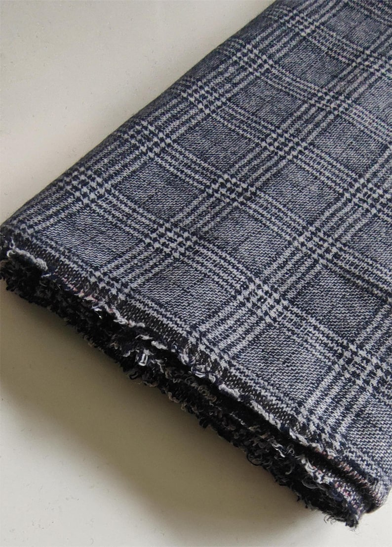 Puede incluir: Una pieza de tela doblada con un patr&oacute;n de cuadros en azul marino oscuro y blanco. El tejido tiene una textura visible y un borde deshilachado y sin rematar a lo largo del pliegue inferior, mostrando la construcci&oacute;n del material.