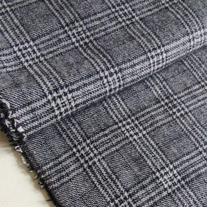 Puede incluir: Una pieza de tela tejida doblada con un patr&oacute;n de cuadros glen en azul marino oscuro y blanco. El textil muestra un tejido texturizado visible con un borde deshilachado en el lateral, destacando el intrincado dise&ntilde;o de cuadr&iacute;cula del material.