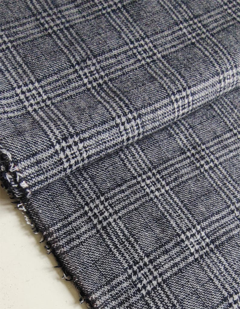 Puede incluir: Una pieza de tela tejida doblada con un patr&oacute;n de cuadros glen en azul marino oscuro y blanco. El textil muestra un tejido texturizado visible con un borde deshilachado en el lateral, destacando el intrincado dise&ntilde;o de cuadr&iacute;cula del material.