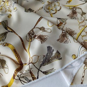 Pode incluir: Um tecido de cor creme com uma estampa repetida de correntes douradas, borlas castanhas e desenhos intrincados de cordas. O material tem uma textura suave e está drapeado para mostrar claramente o padrão na superfície do tecido.
