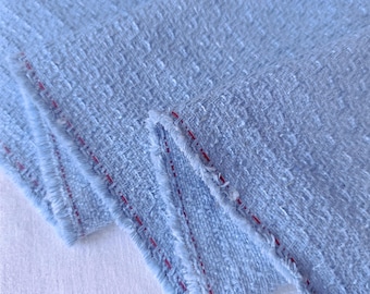 May include: A light blue textured fabric with a woven pattern and frayed edges. The fabric features a distinct red thread detail running along the selvage edge, showcasing a dense, nubby surface texture characteristic of a tweed-style textile.