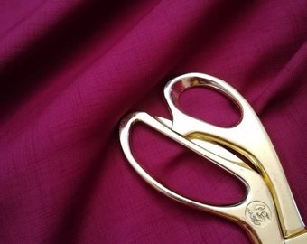 May include: A close-up of a deep burgundy fabric with a pair of gold scissors lying on top. The fabric has a subtle texture and the scissors have a classic design.