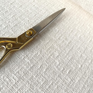 Könnte beinhalten: Eine scharfe Metallschere mit einem polierten goldfarbenen Griff und einer silberfarbenen Klinge. Der Griff ist mit einem kleinen kreisförmigen Emblem und einer zentralen Schraube versehen. Die Schere liegt auf einem weißen Stoff mit Struktur.