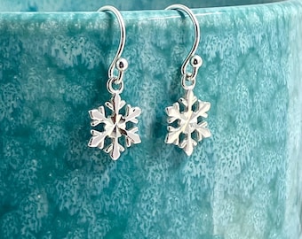 May include: A pair of silver-toned metal earrings featuring a six-pointed snowflake design. Each earring hangs from a curved hook wire, showcasing a polished, reflective surface with intricate, symmetrical cutouts forming the snowflake shape.