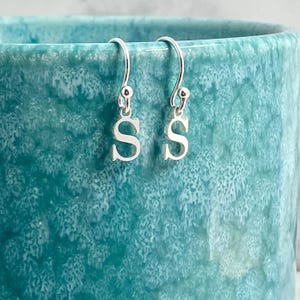 May include: A pair of silver drop earrings, each shaped as the letter "S". The earrings are suspended from a curved hook. The earrings are set against a textured, turquoise background.