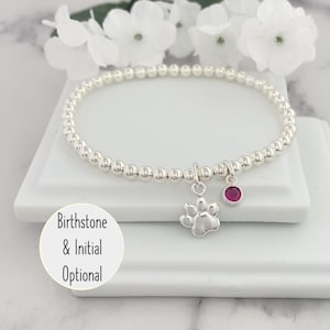 May include: A silver bracelet with a paw print charm and a small red gemstone. The bracelet is on a white surface with the text "Birthstone & Initial Optional".