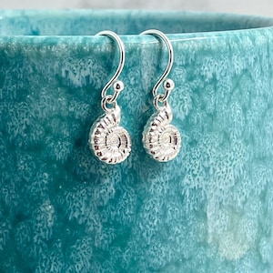 May include: A pair of silver drop earrings featuring a detailed ammonite fossil spiral design. Each earring hangs from a curved silver hook, showcasing the intricate, textured ridges of the shell-like shape in a polished metallic finish.