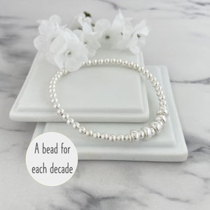 May include: A silver beaded stretch bracelet featuring small round beads and a series of slightly larger, flattened disc-shaped beads. A circular white graphic overlay contains the black text A bead for each decade.