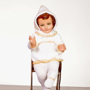 Peut inclure: Une poupée en tricot blanc avec des garnitures dorées est assise sur une chaise en bois. La poupée porte une tenue à capuche en tricot blanc avec des garnitures dorées. La poupée a un visage réaliste avec des cheveux bruns et des yeux bleus.