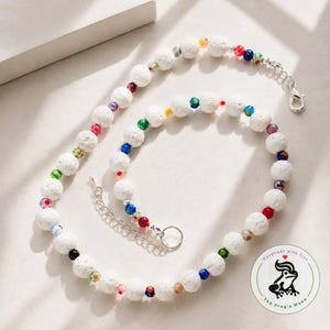 May include: A beaded necklace featuring white porous lava stone beads interspersed with smaller, multicolored glass beads. The necklace includes a silver-toned metal lobster clasp and an adjustable chain extender. A circular logo with a frog graphic is visible.