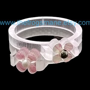 May include: A clear plastic ring with two pink flowers. The flowers have a pearl center and are accented with silver glitter.