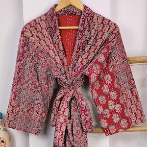 May include: A red and white patterned kimono robe with a tie belt. The robe is hanging on a wooden hanger against a white wall.