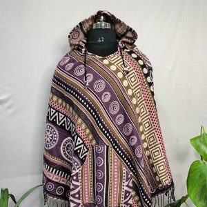 Könnte beinhalten: Ein lila, rosa und braunes Kapuzenponcho mit geometrischem Muster und Fransenbesatz. Das Poncho hat eine Tasche auf der Vorderseite.