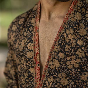 Può includere: Un accappatoio nero con un motivo floreale nei toni dell'oro e del marrone. L'accappatoio ha un bordo rosso e oro lungo i bordi. Il tessuto sembra morbido e fluido, adatto per rilassarsi o come capo decorativo.