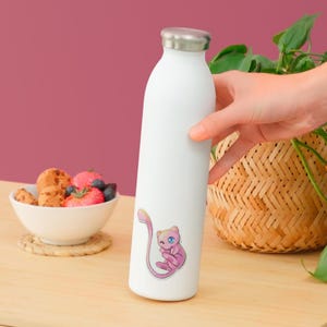 May include: A white cylindrical water bottle with a metallic silver cap, featuring a small, colorful sticker of a pink, cat-like creature with a long tail and blue eyes positioned on the lower front section of the bottle's smooth exterior surface.