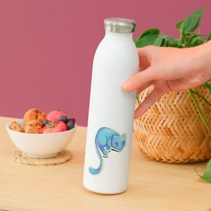 May include: A white stainless steel water bottle featuring a sticker of a stylized blue cat with a long tail. The bottle has a silver-colored metal cap and a smooth, cylindrical body. The cat illustration is rendered in shades of light blue.