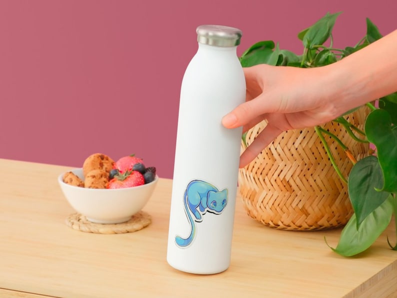 May include: A white stainless steel water bottle featuring a sticker of a stylized blue cat with a long tail. The bottle has a silver-colored metal cap and a smooth, cylindrical body. The cat illustration is rendered in shades of light blue.