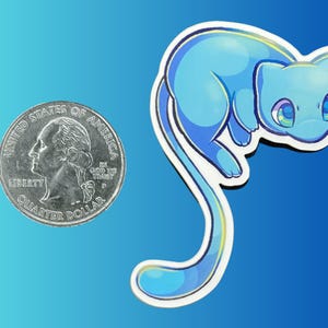 May include: A die-cut sticker featuring a stylized light blue creature with large, expressive eyes and a long, curved tail. The sticker is positioned next to a United States quarter coin for scale, showing the relative size of the illustration.
