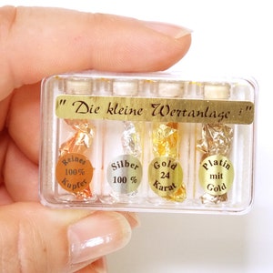 Könnte beinhalten: Ein durchsichtiger Plastikbehälter mit vier kleinen Glasfläschchen, die Gold-, Silber-, Kupfer- und Platinflocken enthalten. Das Etikett auf dem Behälter lautet "Die kleine Wertanlage".