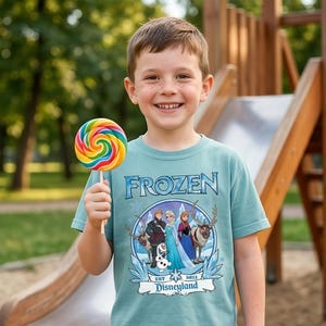 May include: A light blue t-shirt with the word "FROZEN" in a stylized font. The shirt features a graphic of characters from the movie Frozen, including Elsa, Anna, Kristoff, and Olaf, with the text "EST 2013 Disneyland". A colorful lollipop is held in front of the shirt.