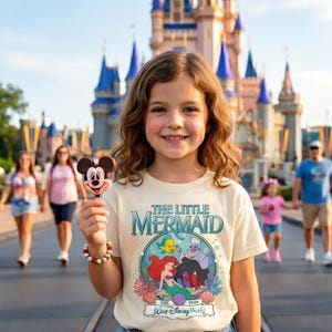 May include: A young person wearing a cream-colored t-shirt featuring "The Little Mermaid" graphic and holding a Mickey Mouse-shaped treat. The shirt has a colorful design with Ariel, Flounder, and Sebastian. The background includes a castle and other people.