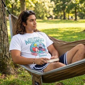 May include: White t-shirt featuring "The Little Mermaid" characters and Disneyland text. The person is wearing the shirt while relaxing in a brown hammock. The person is also wearing dark blue shorts with white stripes.