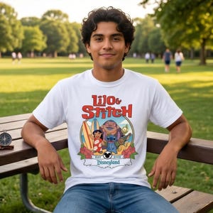 Op de afbeelding: Wit t-shirt met een kleurrijke afbeelding van Lilo & Stitch-personages, surfplanken en bloemen. De tekst "Lilo & Stitch" is in rood en oranje, met "Disneyland" eronder. Het t-shirt wordt gedragen door een persoon die op een parkbank zit.