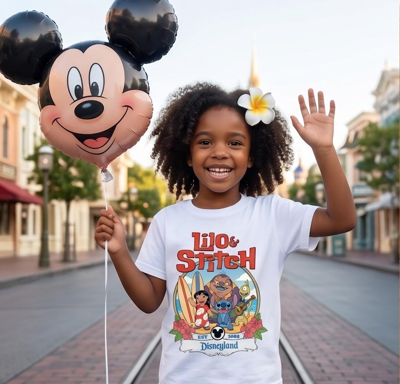 Op de afbeelding: Een jong persoon draagt een wit t-shirt met een kleurrijke "Lilo & Stitch" afbeelding en een Mickey Mouse ballon. Het shirt toont de personages Lilo, Stitch en anderen, met de tekst "Disneyland" eronder. Een bloem zit in hun haar.