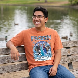 May include: Orange short-sleeved t-shirt featuring the word "FROZEN" in blue, with a graphic of characters from the movie. The shirt also includes the text "EST 2013" and "Walt Disney World". The person is wearing blue jeans.