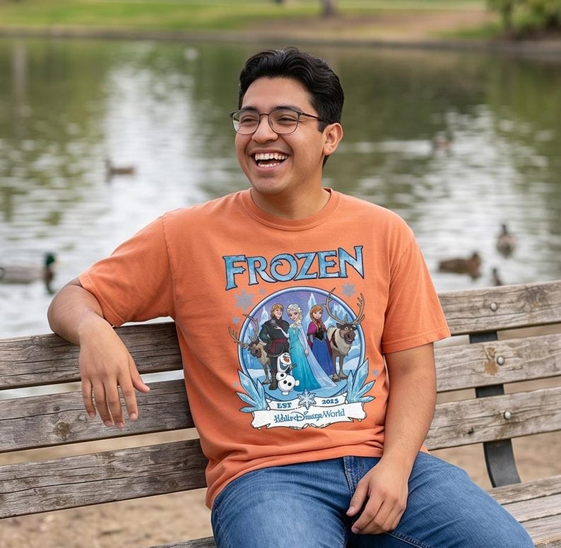 May include: Orange short-sleeved t-shirt featuring the word "FROZEN" in blue, with a graphic of characters from the movie. The shirt also includes the text "EST 2013" and "Walt Disney World". The person is wearing blue jeans.