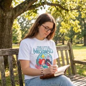 May include: A white t-shirt featuring the text "THE LITTLE MERMAID" in teal and a colorful graphic of Ariel, Flounder, Sebastian, and Ursula. The shirt is worn by a person sitting on a wooden bench outdoors, writing in a brown leather-bound journal.