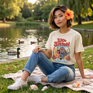 Op de afbeelding: Een cr&egrave;mekleurig T-shirt met de woorden "Lilo & Stitch" in het rood en een afbeelding van de personages. Het shirt wordt gedragen met een blauwe spijkerbroek en witte sneakers. De foto is buiten genomen met een meer en bomen op de achtergrond.