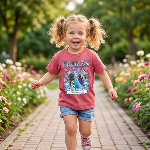 May include: A young child wearing a red t-shirt with the word "FROZEN" and a graphic of characters from the movie. The child is also wearing blue denim shorts and pink sneakers. The child is running on a brick pathway.