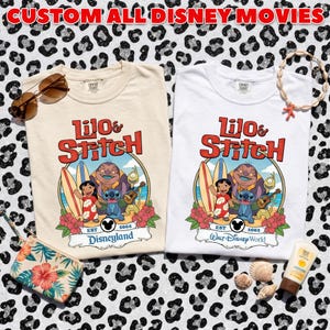 Op de afbeelding: Twee T-shirts, &eacute;&eacute;n cr&egrave;me en &eacute;&eacute;n wit, met een kleurrijke afbeelding van Lilo & Stitch met surfplanken en bloemen. Het cr&egrave;mekleurige shirt zegt "Disneyland" en het witte shirt zegt "Walt Disney World." De tekst "CUSTOM ALL DISNEY MOVIES" staat bovenaan.