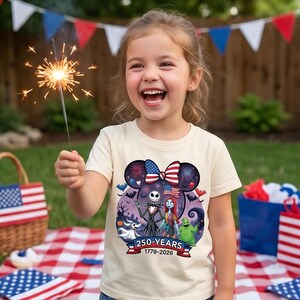 May include: A cream-coloured short-sleeve t-shirt featuring a graphic of Jack Skellington, Sally, Zero, and Oogie Boogie. The design includes a Mickey-shaped silhouette with a patriotic bow, fireworks, and the text 250-YEARS 1778-2026.