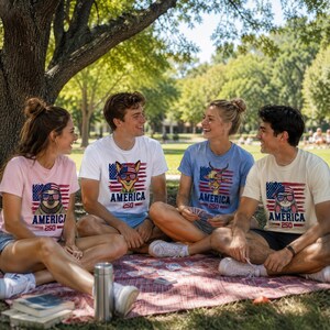 May include: Four people sitting on a picnic blanket, wearing t-shirts with patriotic animal graphics and the word "AMERICA". The shirts are in various colors: pink, white, blue, and cream. The graphics include a sloth, dog, goat, and rabbit, all wearing sunglasses.