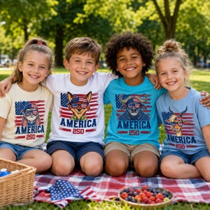 May include: Four children wearing patriotic t-shirts with animal graphics and the word "AMERICA" in red and blue. The shirts feature a bunny, fox, bull, and goat, each with sunglasses and a flag design. They are sitting on a red and white checkered blanket.