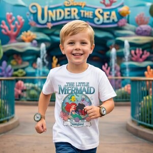 May include: A young child wearing a white t-shirt featuring "The Little Mermaid" characters and the Disneyland logo. The shirt has a colorful graphic of Ariel, Flounder, and Ursula. The child is smiling and wearing a watch on each wrist.