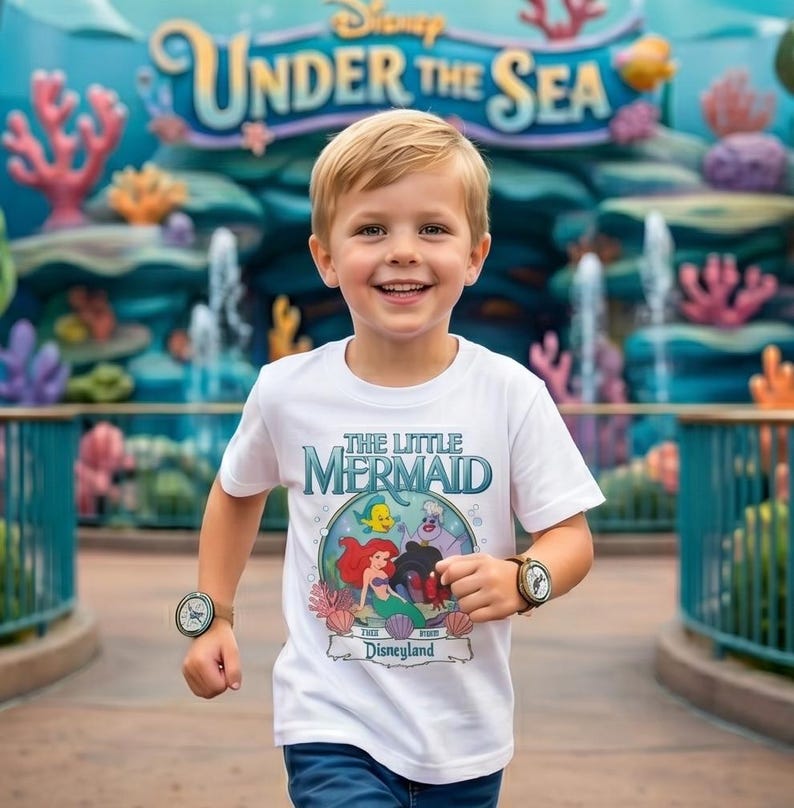 May include: A young child wearing a white t-shirt featuring "The Little Mermaid" characters and the Disneyland logo. The shirt has a colorful graphic of Ariel, Flounder, and Ursula. The child is smiling and wearing a watch on each wrist.