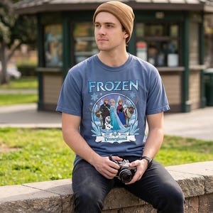 May include: A blue t-shirt with the word "FROZEN" in white and blue lettering. The shirt features a circular graphic with characters from the movie Frozen, including Elsa, Anna, Kristoff, Sven, and Olaf. The text "EST 2013 Disneyland" is also present.