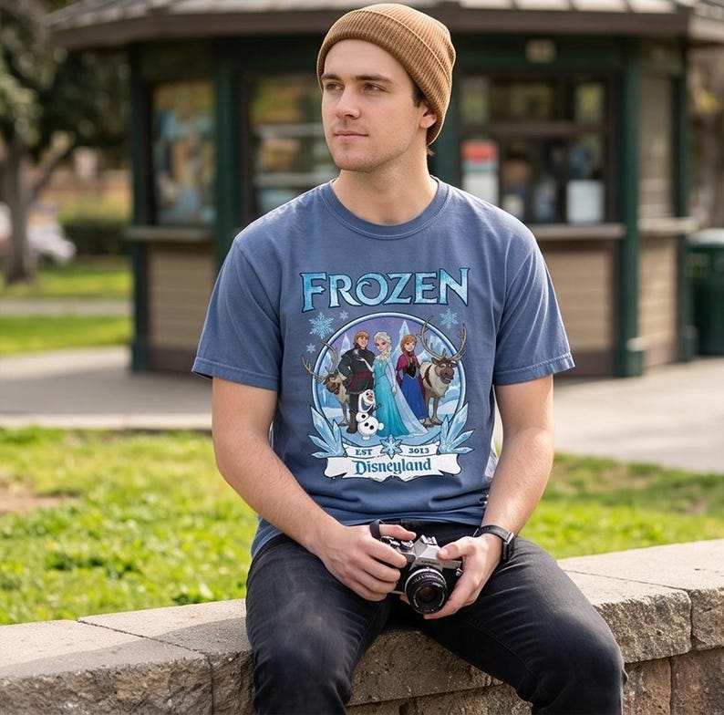 May include: A blue t-shirt with the word "FROZEN" in white and blue lettering. The shirt features a circular graphic with characters from the movie Frozen, including Elsa, Anna, Kristoff, Sven, and Olaf. The text "EST 2013 Disneyland" is also present.