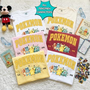 May include: A collection of six crew neck t-shirts in various colors, including white, pink, yellow, and burgundy. Each shirt features the word "POKEMON" in a yellow collegiate font, along with a graphic of several Pokemon characters. The text "Est. 1998" is also printed on each shirt.