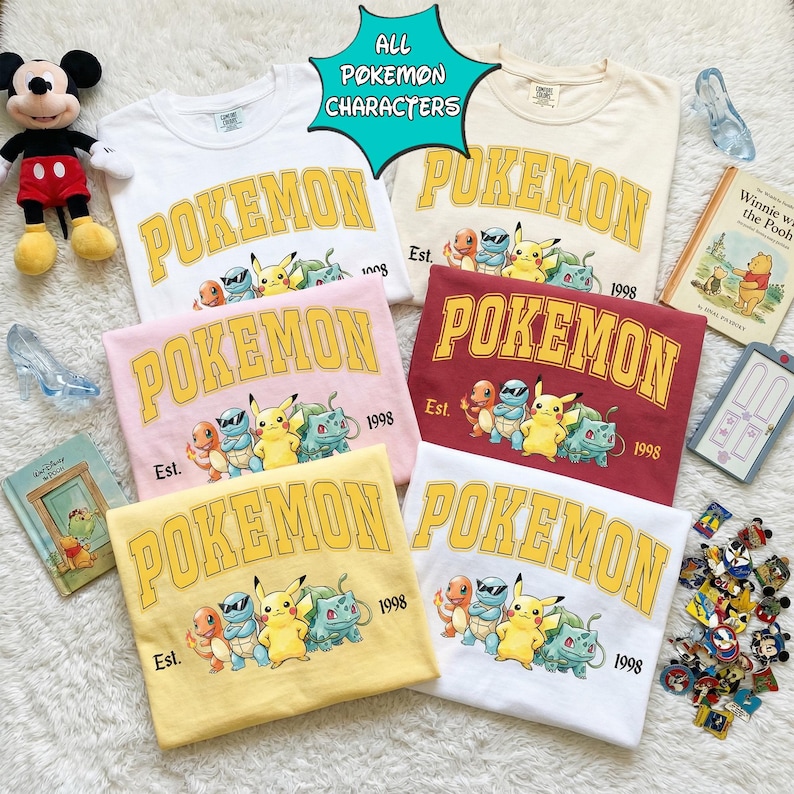 May include: A collection of six crew neck t-shirts in various colors, including white, pink, yellow, and burgundy. Each shirt features the word "POKEMON" in a yellow collegiate font, along with a graphic of several Pokemon characters. The text "Est. 1998" is also printed on each shirt.