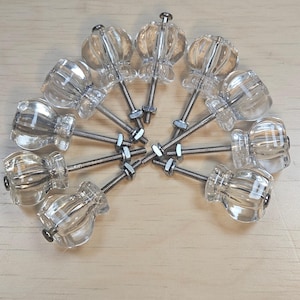 May include: A collection of clear glass cabinet knobs with a decorative, fluted design. Each knob has a silver-colored screw and nut for installation. The knobs are arranged in a circular pattern on a light wood surface.