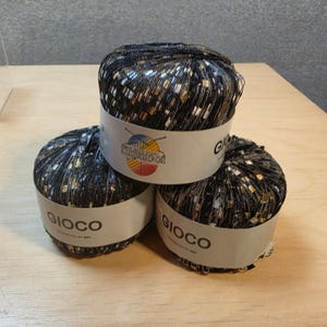 May include: Three balls of Gioco yarn featuring a black base with metallic gold and silver flecks. Each ball is wrapped in a white paper band with the text Gioco and a logo depicting a ball of yarn with knitting needles. The yarn has a shiny texture.
